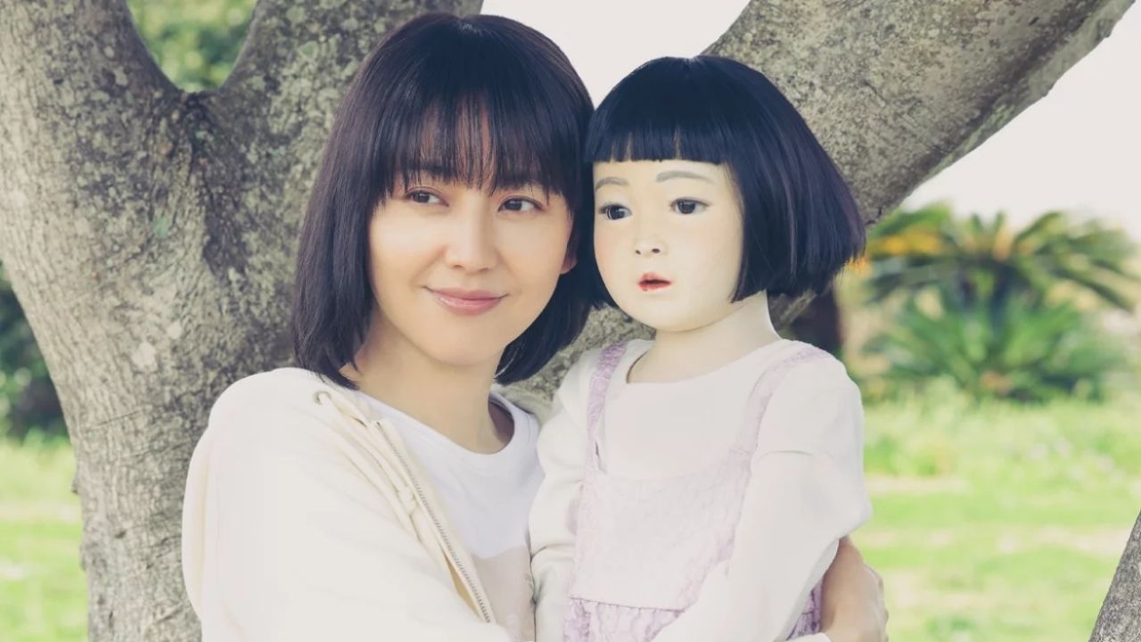 Uma mulher de cabelos escuros e curtos segura uma boneca de aparência realista com rosto pintado e cabelo preto liso, também curto. Ambas estão vestidas com roupas claras, e a cena ocorre ao ar livre, com uma árvore robusta ao fundo e gramado verde ao redor, sugerindo um ambiente de parque em um dia ensolarado.