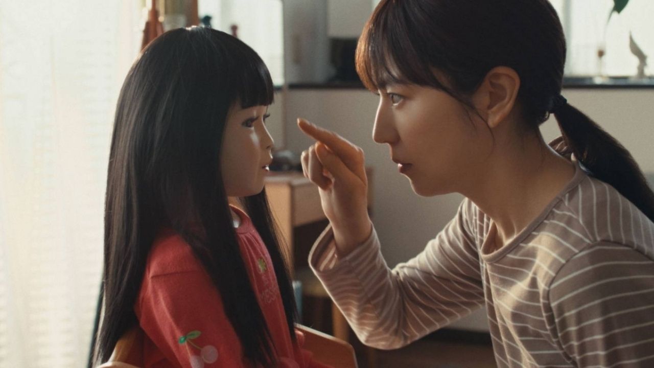 Uma mulher de cabelos longos e escuros, presos em um rabo de cavalo, usa uma blusa listrada bege e aponta o dedo para o rosto de uma criança com cabelos longos e pretos, vestida com uma blusa vermelha decorada com cerejas. Elas estão sentadas em uma mesa dentro de uma casa iluminada por luz natural que entra pelas janelas com cortinas claras, criando uma atmosfera aconchegante.