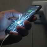 Mão segurando um celular enquanto um cabo de carregador danificado solta faíscas de choque elétrico.