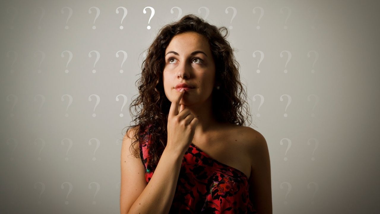 Uma mulher de cabelos cacheados e escuros, vestindo um vestido vermelho com estampa floral, está com a mão no queixo em pose pensativa. O fundo é cinza claro com vários ícones de ponto de interrogação brancos, sugerindo um tema de curiosidade ou reflexão.
